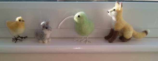 A needle felted yellow chicken, grey rabbit, green kiwi and yellow fox sitting on a ledge
